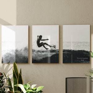 Może przedstawiać: Trzy czarno-białe, oprawione wydruki przedstawiające surferów na fali. Środkowy wydruk przedstawia surferów w akcji, podczas gdy wydruki po lewej i prawej stronie pokazują falę z różnych perspektyw. Prawy wydruk zawiera tekst "Praia da Conceição, Brasil".