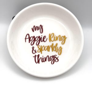 Puede incluir: Un plato de cerámica blanco con el texto "my Aggie Ring & Sparkly things" en letras granate y doradas. El plato es redondo con un borde ligeramente elevado, perfecto para guardar pequeñas joyas.