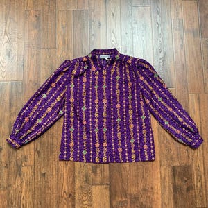 May include: A long-sleeved purple blouse with a high collar. The blouse features a vertical striped pattern with floral and geometric accents in gold and green. The fabric has a slight sheen, and the sleeves are gathered at the cuffs.