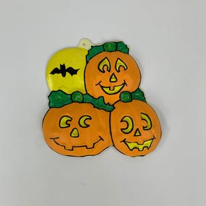 May include: A Halloween decoration featuring three orange jack-o'-lanterns with smiling faces and green leaves. A yellow moon with a black bat silhouette is also present. The decoration is made of a lightweight material.
