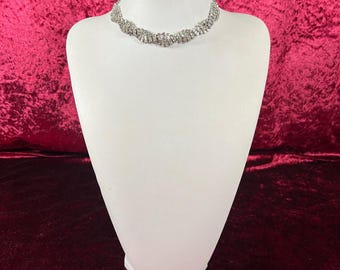 May include: A silver-toned rhinestone necklace displayed on a white mannequin bust. The necklace is a choker style, featuring multiple rows of sparkling rhinestones. The background is a deep red velvet.