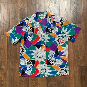 May include: A short-sleeved button-up shirt with a vibrant geometric pattern. The shirt features a collage of colors including red, orange, green, purple, and blue, with white and black paisley and sun designs. The shirt is made of a lightweight fabric.