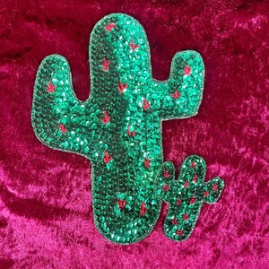 May include: Two sequined cactus patches. The larger cactus is green with red accents, and the smaller one is a miniature version of the larger one. The patches are on a deep pink velvet background.
