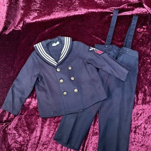 May include: A navy blue sailor-style outfit consisting of a double-breasted jacket with gold-tone buttons, a wide collar with three white stripes, and long trousers with attached suspenders. A patch with a red and white eagle emblem is on the sleeve.