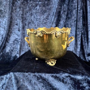 May include: A gold-toned metal bowl featuring a scalloped rim with decorative leaf-like accents, two small side handles, and three ornate feet. The bowl has a smooth, polished surface and a rounded body shape with intricate detailing along the top edge.