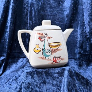 May include: A cream-colored ceramic teapot with a square-shaped body and a matching lid. The exterior features a colorful graphic print depicting a tall blue vase, a yellow bowl, and two smaller bowls in red and yellow with geometric patterns.