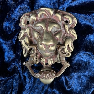 May include: A brass lion head door knocker featuring a detailed mane and a hinged ring held in the lion's mouth. The metal has a weathered, aged finish with visible dark patina in the recessed areas of the casting, including two mounting holes.