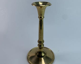 May include: A brass candlestick holder with a round base, a slender stem, and a flared top. The candlestick is a warm gold color and has a classic design. The base is approximately 3 inches in diameter.