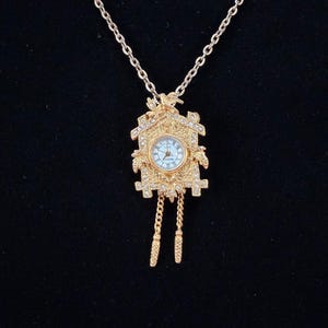 May include: Gold-toned necklace featuring a miniature cuckoo clock pendant. The pendant has a light blue clock face, decorative crystals, and dangling chain accents. The necklace is displayed against a black background.