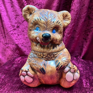 May include: A brown ceramic bear cookie jar with blue eyes, a black nose, and a red tongue. The bear has a lid and is sitting with its paws out. The jar is painted with brown and tan tones.