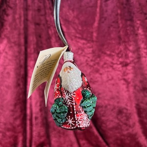 May include: A Christmas ornament shaped like Santa Claus. The ornament features a red coat with white snowflake designs and green glittery tree-shaped mittens. Santa has a white beard and a silver cap. A tag is attached to the ornament.