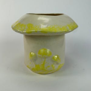 May include: A ceramic mushroom-shaped vase with a cream-colored body and a yellow-accented cap. The vase features three small, yellow mushroom details on the front. The interior is a darker, earthy tone.