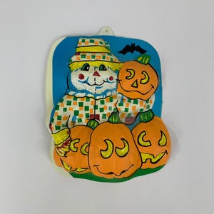 May include: A vintage Halloween decoration featuring a scarecrow and jack-o'-lanterns. The scarecrow wears a plaid hat and shirt, with a smiling face. Three orange pumpkins with smiling faces are at the bottom, with a bat silhouette above.