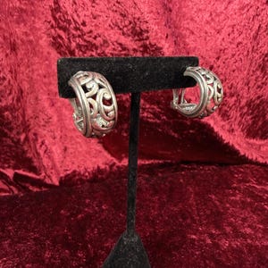 May include: A pair of silver hoop earrings with an open filigree design. The earrings are displayed on a black velvet earring stand against a red velvet backdrop. The earrings have a hinged closure.