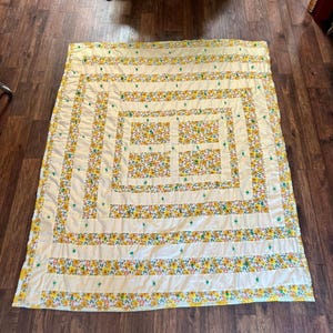 May include: A rectangular quilt with a floral pattern in yellow, orange, and green on a cream background. The quilt features a striped design with a central square and is laid out on a wooden floor.