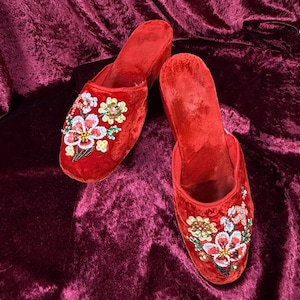 May include: A pair of red velvet slippers with floral embroidery. The slippers feature a closed-toe design and are adorned with colorful beads and sequins in a flower pattern. The background is a deep red velvet fabric.