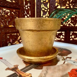 May include: A gold-colored flower pot with a matching saucer. The pot has a textured surface and a classic shape. The pot and saucer are sitting on a tray with a musical theme.