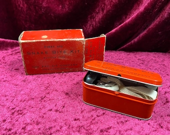 Könnte beinhalten: Ein rotes Vintage-Schlangenbiss-Kit mit dem Text "First Aid Snake Bite Kit" auf der Box. Das Kit enthält einen roten Metallbehälter mit medizinischen Utensilien. Auf der Box ist auch die Pac-Kit Company aufgedruckt.