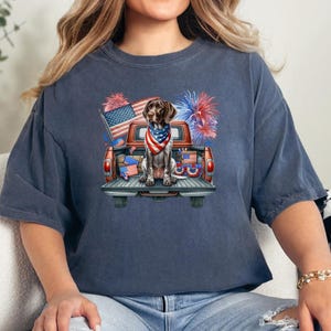 May include: Navy blue t-shirt featuring a German Shorthaired Pointer dog wearing a patriotic bandana, sitting in the back of a red pickup truck decorated with American flags and fireworks. The design celebrates Independence Day.
