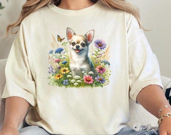 May include: A white t-shirt with a graphic of a chihuahua dog sitting in a field of flowers. The dog is brown and white with a butterfly flying above it.