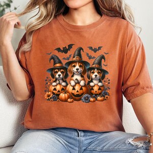 May include: Orange t-shirt featuring a design of three beagle puppies wearing witch hats and holding jack-o'-lanterns. Bats are flying above the puppies.