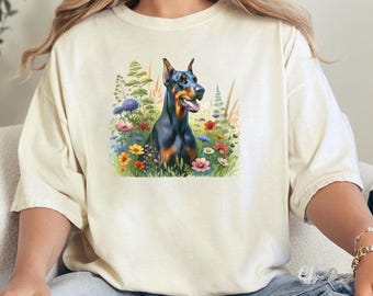 May include: A white t-shirt with a black Doberman Pinscher dog standing in a field of flowers. The dog is looking to the left of the image. The flowers are in shades of pink, purple, yellow, and blue.
