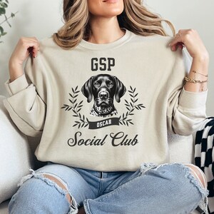 May include: A beige sweatshirt with a black and white German Shorthaired Pointer dog illustration. The text "GSP" is above the dog and "Oscar" is below the dog. The text "Social Club" is below the dog's image.