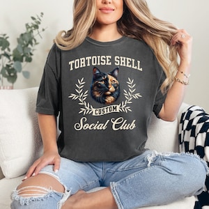 May include: A gray t-shirt with the text "Tortoise Shell Custom Social Club" and a picture of a calico cat.