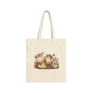 Könnte beinhalten: Eine cremefarbene Stofftasche mit einer zentralen grafischen Illustration von vier kleinen, lächelnden Geistern, umgeben von bunten Wildblumen, grünem Blattwerk und orangefarbenen Schmetterlingen. Die Geister sind mit Blumenkränzen und Hüten geschmückt.