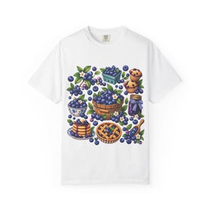 Op de afbeelding: Een wit t-shirt met een kleurrijke grafische print van bosbessen. Het ontwerp bevat een houten mand met bosbessen, een kom met bessen, een bosbessentaart, muffins, een pot jam met een blauw geruit deksel en losse bosbessen.