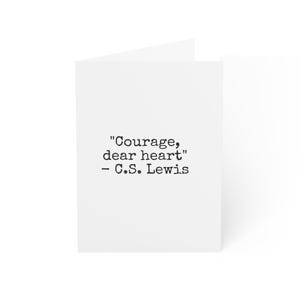 May include: A white greeting card featuring black typewriter-style text centered on the front. The text reads, "Courage, dear heart" followed by the attribution "- C.S. Lewis" on the line below. The card is folded and stands upright.