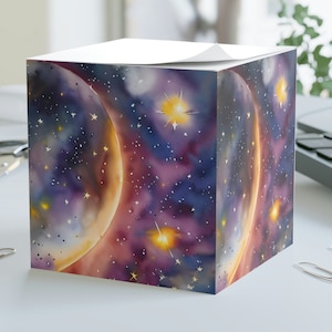 May include: A cube-shaped notepad with a colorful galaxy design. The design features a crescent moon, stars, and nebulae in shades of purple, blue, orange, and yellow.