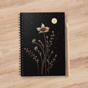 Puede incluir: Una libreta espiral negra con un diseño floral dorado y una luna creciente en la portada. El diseño presenta una flor grande con flores y hojas más pequeñas.