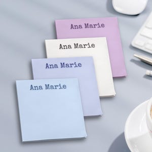 May include: Four square sticky notes in pastel colors: light blue, lavender, white, and pale blue. Each note has the name "Ana Marie" printed in a typewriter-style font. The notes are stacked at an angle, with a keyboard and mouse in the background.