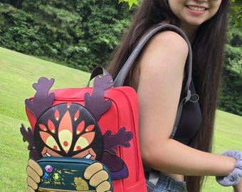 May include: A red backpack with a cartoon character design. The character has a dark face with multiple orange eyes and purple antlers. The backpack has a front pocket with a green and gold design. The straps are gray.