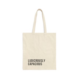 May include: A cream-colored tote bag with the words "LUDICROUSLY CAPACIOUS" printed in black. The bag has two handles and is made of a sturdy fabric, suitable for carrying groceries, books, or other items.