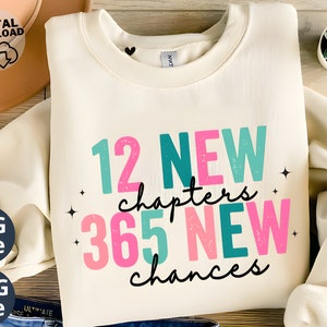 May include: A white sweatshirt with the text "12 NEW chapters 365 NEW chances" in pink, teal, and turquoise lettering with a distressed effect.