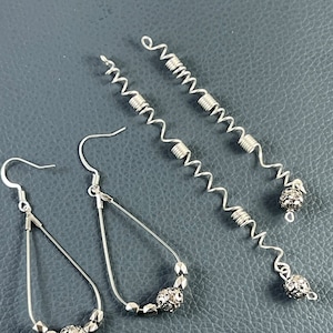 Puede incluir: Tres pares de pendientes de plata. Dos pares presentan una forma de lágrima con un diseño de cuentas y una bola de plata en el extremo. El otro par presenta un alambre largo, plateado y en espiral con una bola de plata en el extremo.