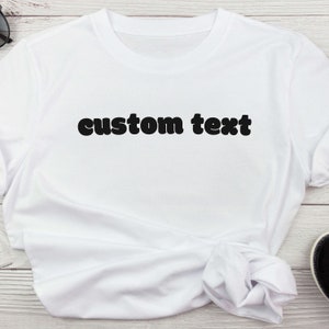 Puede incluir: Camiseta blanca con la frase "custom text" impresa en letras negras y en negrita en el pecho. La camiseta está hecha de una tela suave y ligera y tiene un cuello redondo clásico.