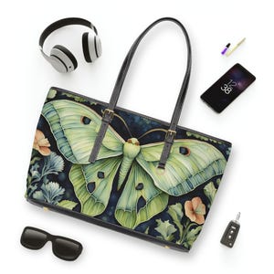 Può includere: Una borsa tote con una grande e dettagliata illustrazione di una falena verde su uno sfondo blu scuro con accenti floreali. La borsa ha manici e finiture nere, con accessori intorno.