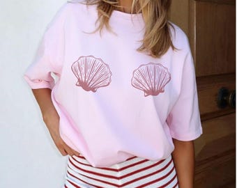 Può includere: Una maglietta rosa chiaro con due disegni di conchiglie in una tonalità rosa più scura. È abbinata a pantaloncini a righe rosse e bianche. L'insieme evoca un tema estivo o balneare.