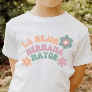 Puede incluir: Camiseta blanca con el texto "LA MEJOR HERMANA MAYOR" en un arco&iacute;ris de colores con flores a cada lado.