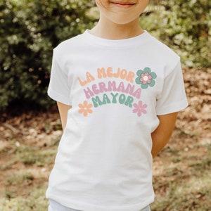 Puede incluir: Camiseta blanca con un gr&aacute;fico de arco&iacute;ris que dice "La mejor hermana mayor" con flores a cada lado.