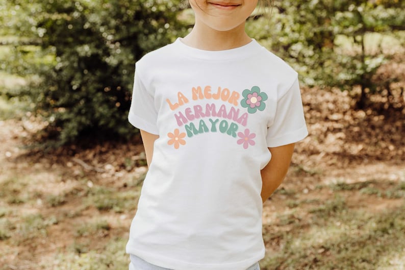 Puede incluir: Camiseta blanca con un gr&aacute;fico de arco&iacute;ris que dice "La mejor hermana mayor" con flores a cada lado.