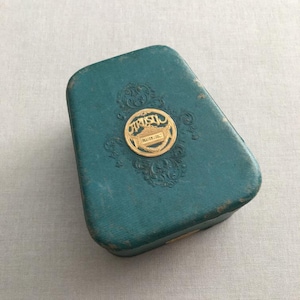 Può includere: Una scatola verde turchese con un'etichetta dorata in rilievo che recita "ARISTOL  PARFUM DOUCE".