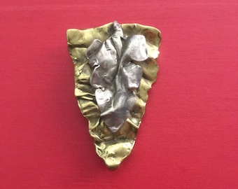 Könnte beinhalten: Handgefertigte Metallbrosche in Blattform. Die Brosche hat eine Kombination aus Silber- und Goldtönen mit strukturierter Oberfläche. Das abstrakte, organische Design ist vor rotem Hintergrund platziert.