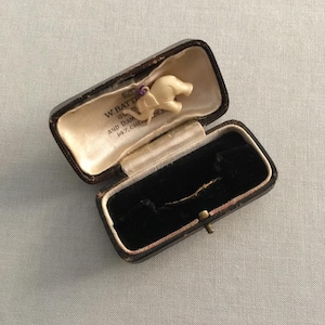 May include: An open antique jewelry box with a small ivory-colored elephant figurine inside. The box is dark brown with a black velvet interior and a cream-colored satin lining. The text "W. BATTY & CO. AND DIAM. 147. CHEAPSIDE" is visible.