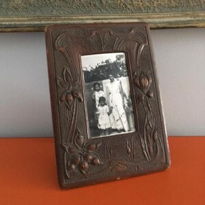 May include: A dark brown wooden picture frame with an ornate carved design featuring stylised flowers and leaves. The frame holds a black and white photograph of three people.
