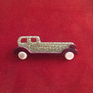 Puede incluir: Un broche con forma de coche vintage con un cuerpo estampado en blanco y negro y detalles burdeos. El coche tiene ruedas blancas nacaradas.