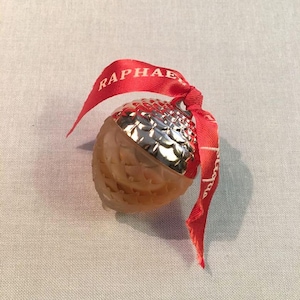 May include: An acorn-shaped perfume bottle with a silver cap and a red ribbon. The bottle is a light brown color and has a textured surface. The red ribbon has the word "Raphael" printed on it.